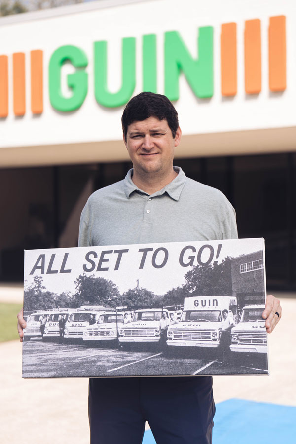 A person holding a vintage poster of Guin company vans, with a Guin sign in the background.